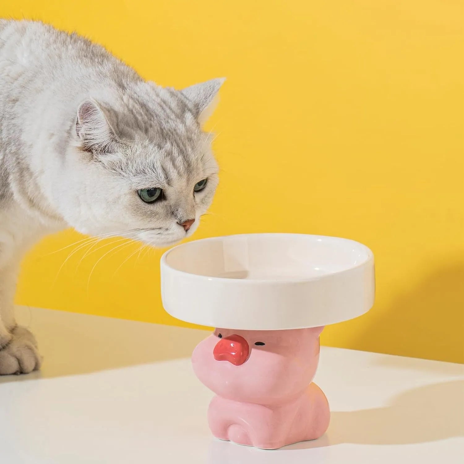 Raised ceramic cat bowl with animal design in pink, yellow, or white on a wooden floor.