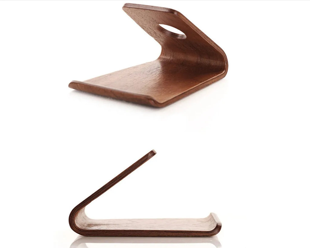 Handmade solid wood iPhone stand in walnut finish on a desk, holding a phone with a charging cable routed through the cable channel.