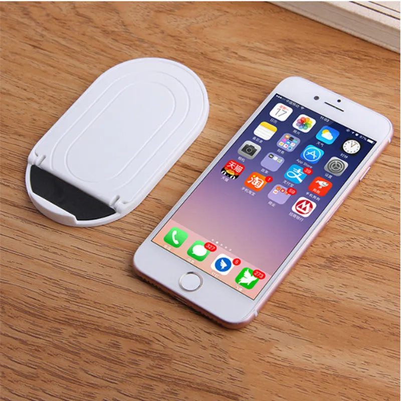 A lazy bracket style mobile phone stand made of plastic, shown in multiple colour options on a wooden desk.