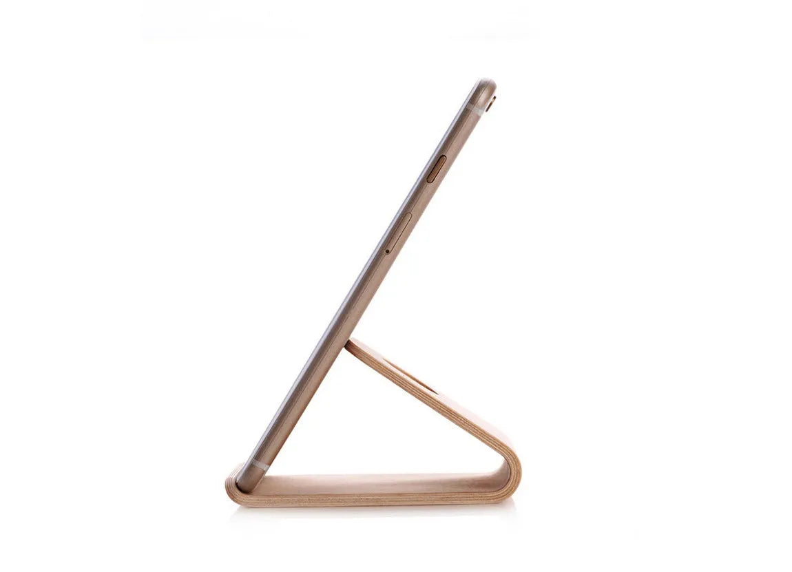 Handmade solid wood iPhone stand in walnut finish on a desk, holding a phone with a charging cable routed through the cable channel.