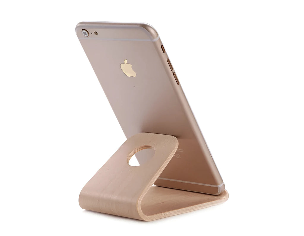 Handmade solid wood iPhone stand in walnut finish on a desk, holding a phone with a charging cable routed through the cable channel.