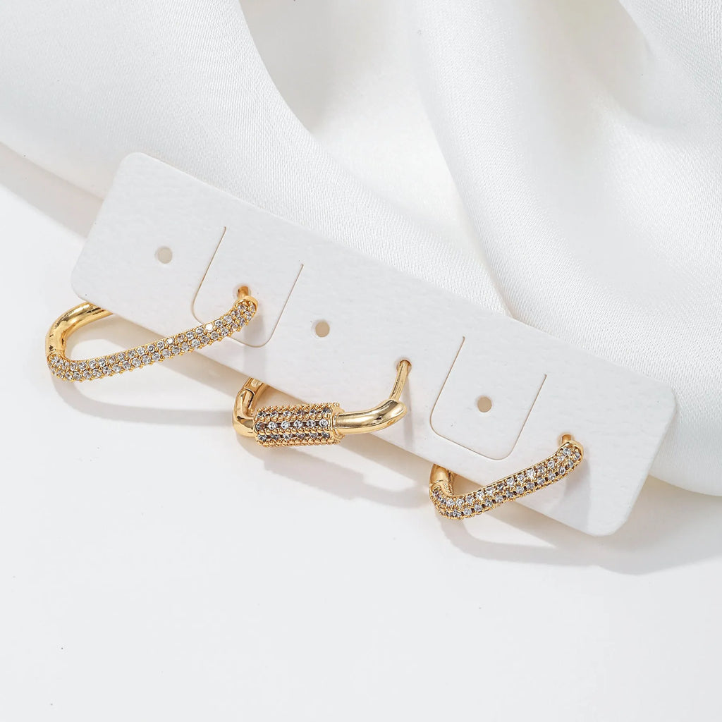 Three geometric square ear clips with inlaid zircon details in gold and white finishes on a grey background.