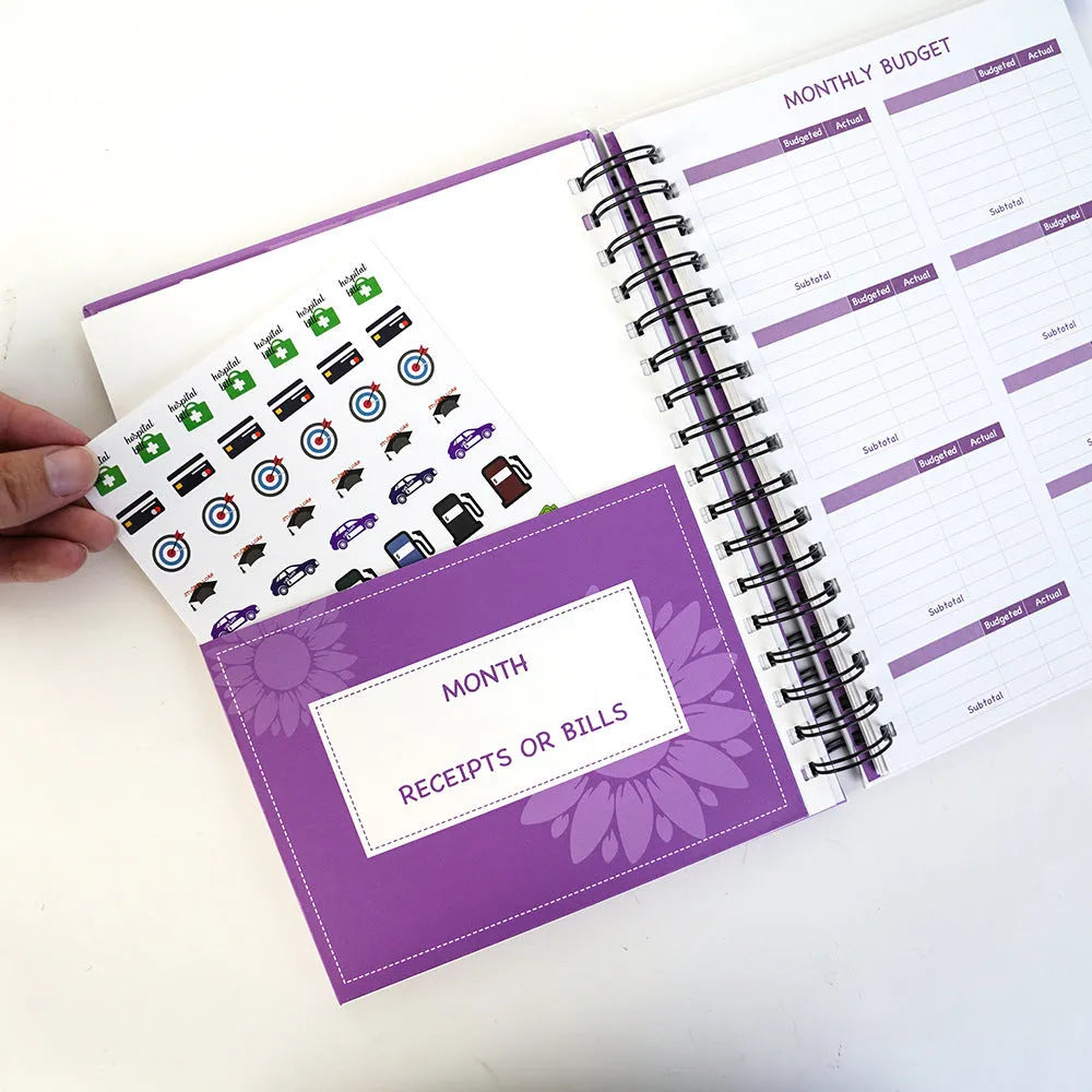 Purple coil-bound budget notebook open showing monthly planning pages with financial stickers and paper clips