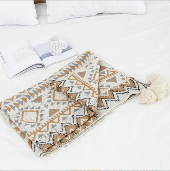 Beige and orange bohemian-style acrylic sofa blanket with tassel fringe on a grey sofa.