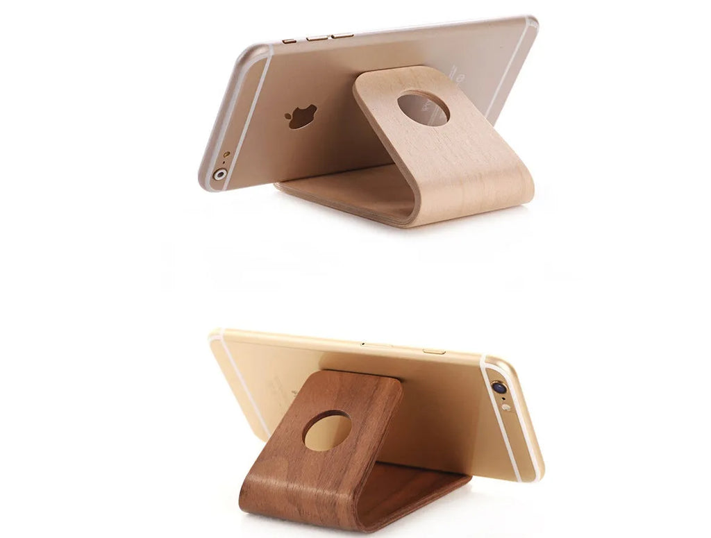 Handmade solid wood iPhone stand in walnut finish on a desk, holding a phone with a charging cable routed through the cable channel.
