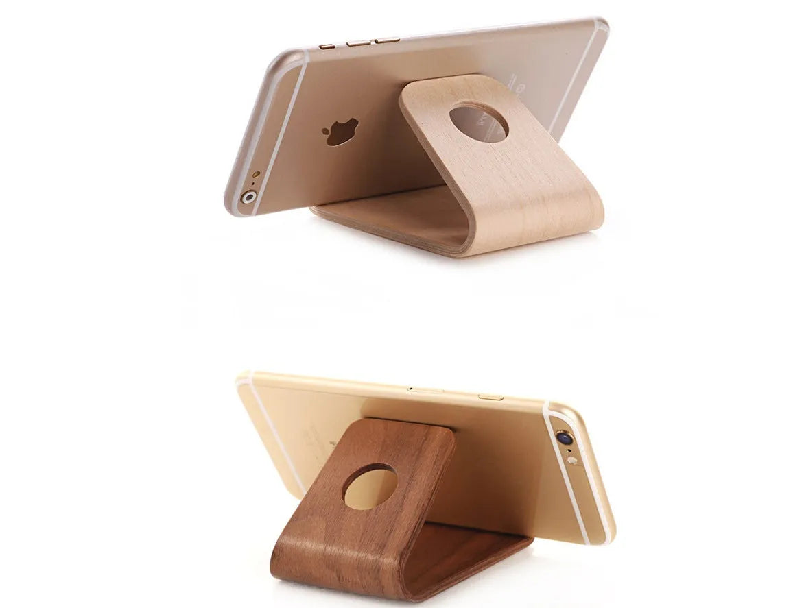 Handmade solid wood iPhone stand in walnut finish on a desk, holding a phone with a charging cable routed through the cable channel.