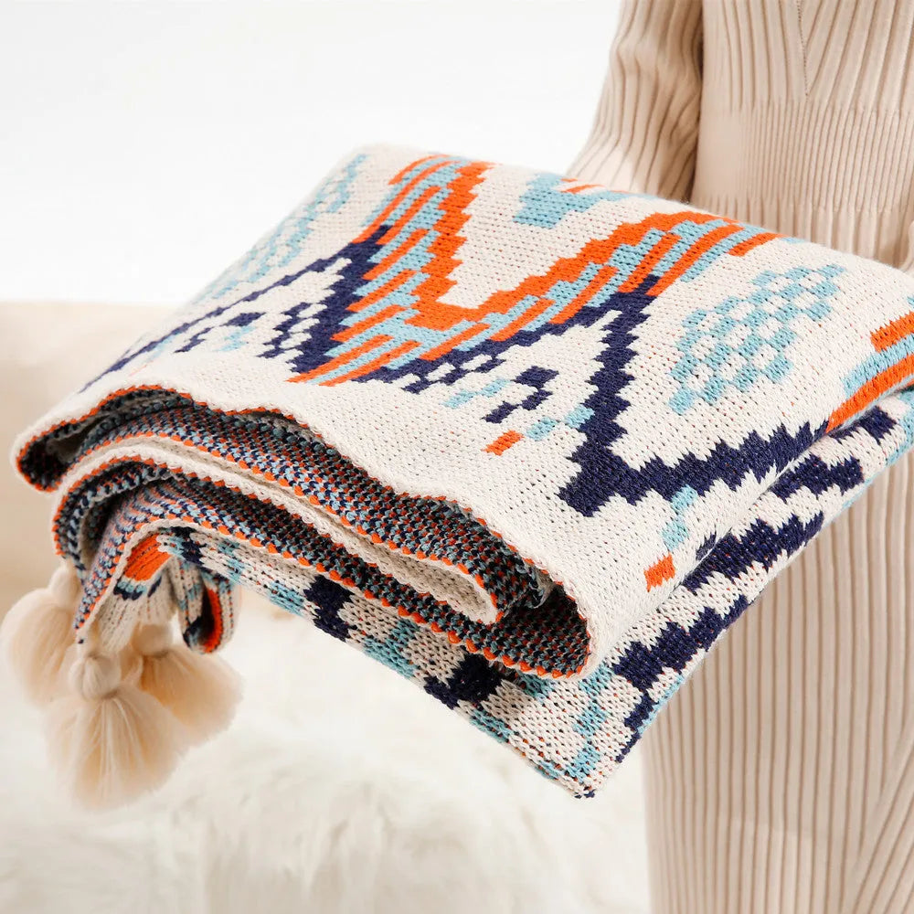 Beige and orange bohemian-style acrylic sofa blanket with tassel fringe on a grey sofa.