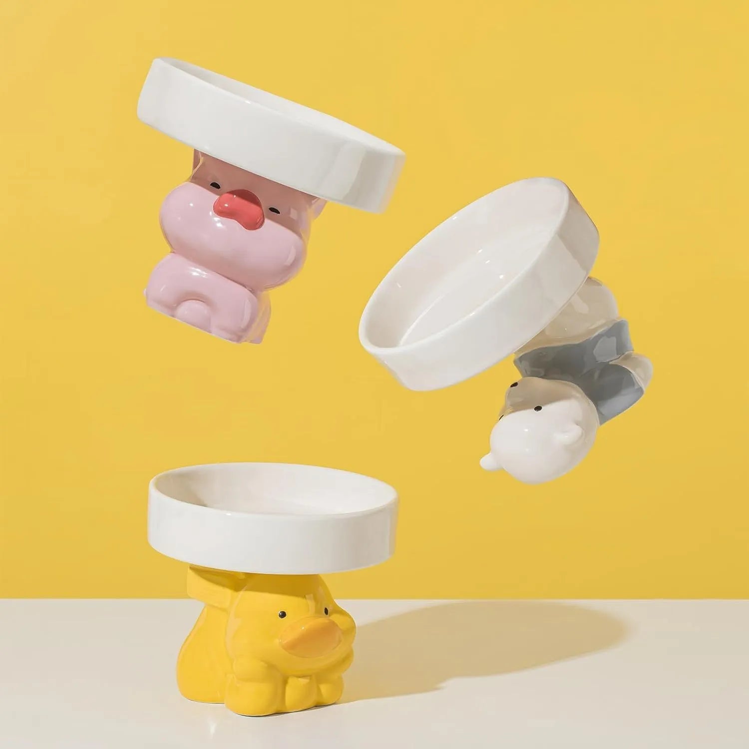 Raised ceramic cat bowl with animal design in pink, yellow, or white on a wooden floor.
