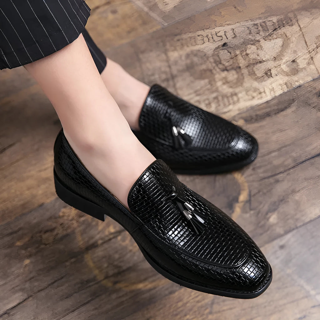 Men's black pointed toe slip on loafers with a tassel detail on the front, shown on a plain background.