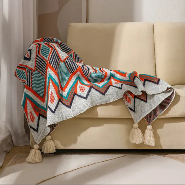 Beige and orange bohemian-style acrylic sofa blanket with tassel fringe on a grey sofa.